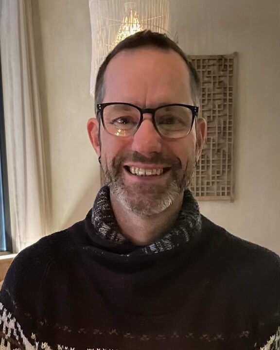 Portrait of Rich Bothe, smiling, wearing glasses and a dark patterned sweater